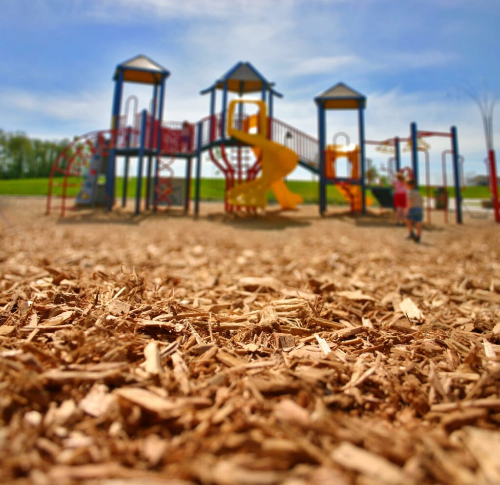 Ground view close-up shredded wood chip material with children approaching composite play structure in background